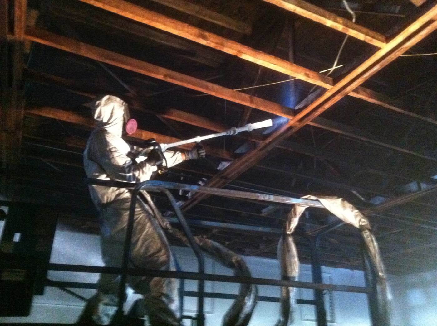 Technician in protective suit dry ice blasting structural steel roof framing inside a warehouse